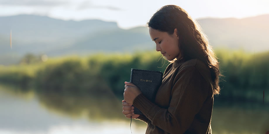 A young women near a lake holding a bible in reflection and thanks.