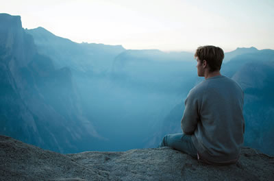 A man sitting on the edge of a cliff staring at the nearby mountains